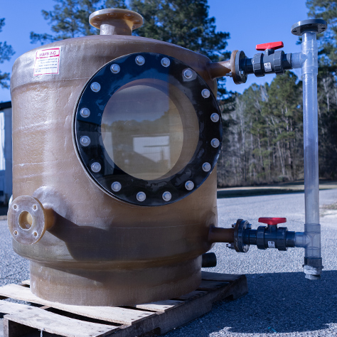 mermade-filter-commercial-pool-surge-tank-assembly-fiberglass-474-474 This is an image of a Mer-Made Filter fiberglass commercial pool surge tank on a wooden pallet. The tank is tan-colored fiberglass with an outlet on the lower left side and a large manway with clear cover. The right side has two outlets that connect to a clear tube with shut off valves at each end.